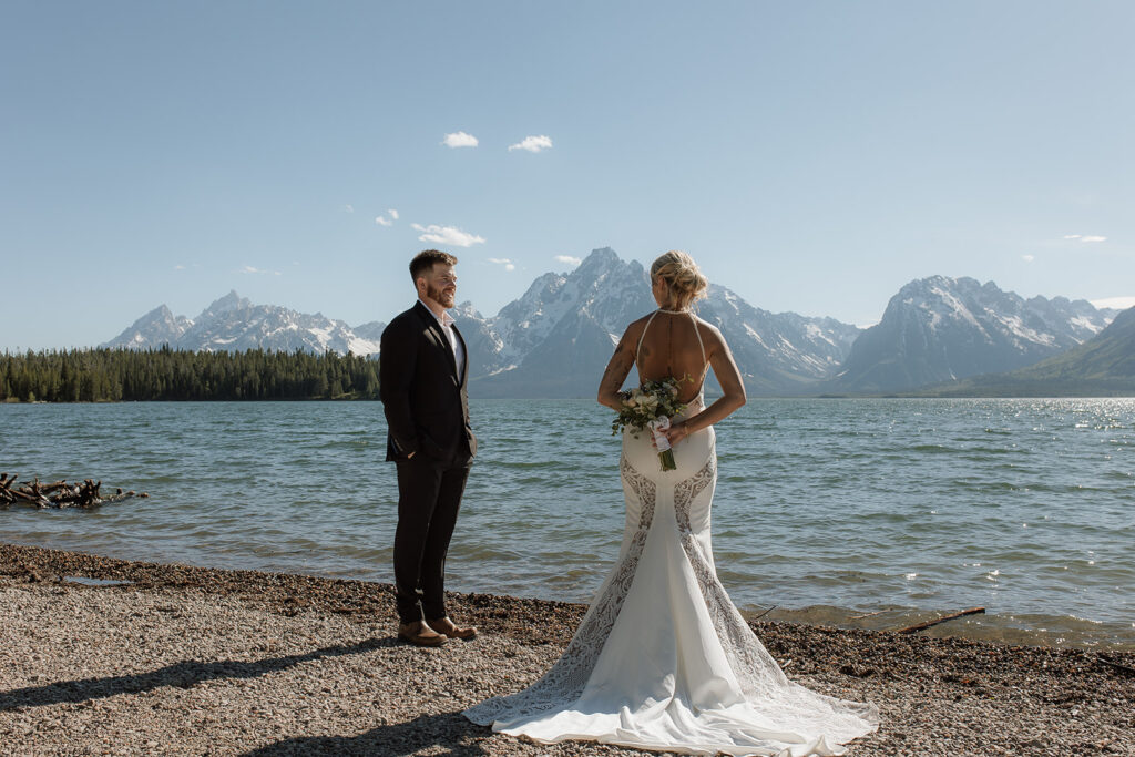 A couple stands on a rocky beach with a mountainous lake backdrop. The man embraces the woman from behind as they both look into the distance.