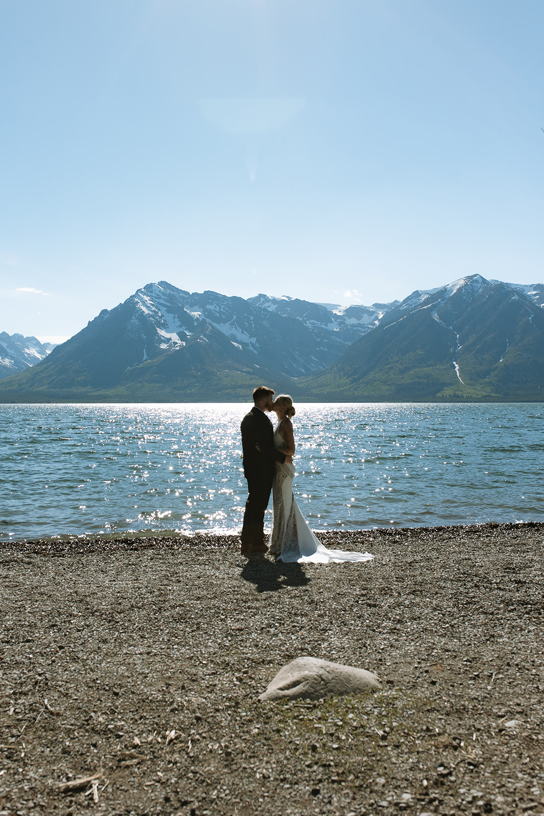   A couple dressed in formal attire stands together on a pebble shore in front of a lake, with snow-capped mountains in the background under a clear blue sky.