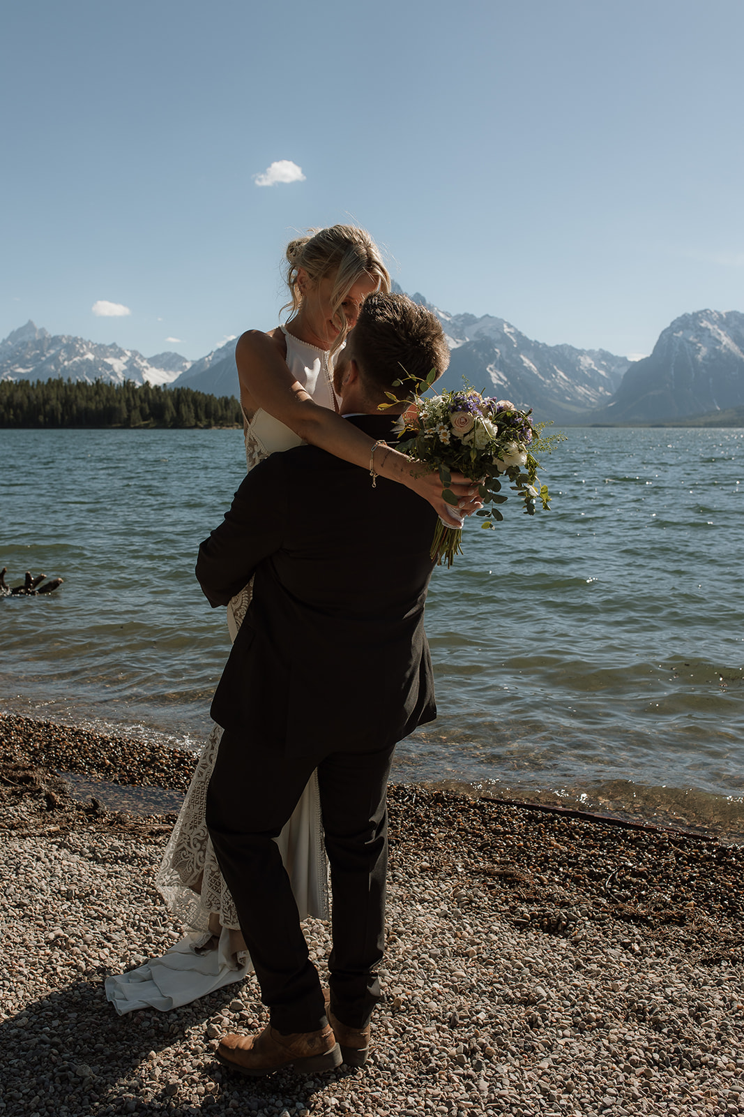 A couple dressed in formal attire stands together on a pebble shore in front of a lake, with snow-capped mountains in the background under a clear blue sky.