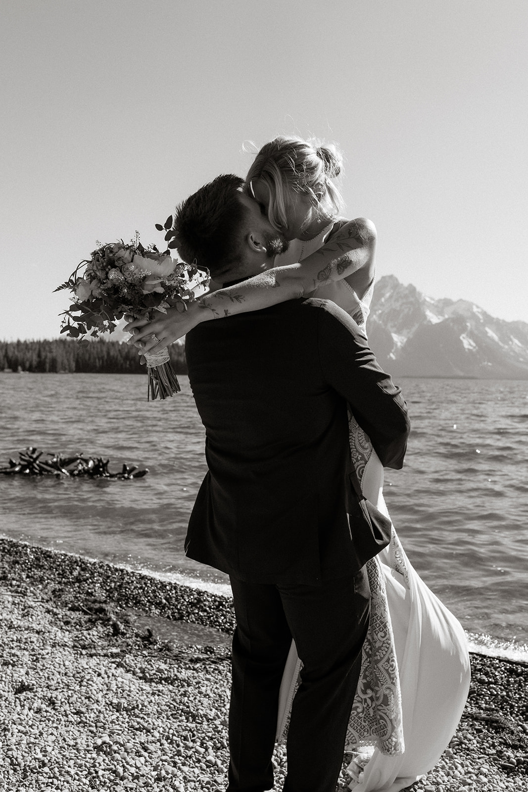 A bride and groom share a kiss by a lake, with mountains in the background. The groom lifts the bride who holds a bouquet.