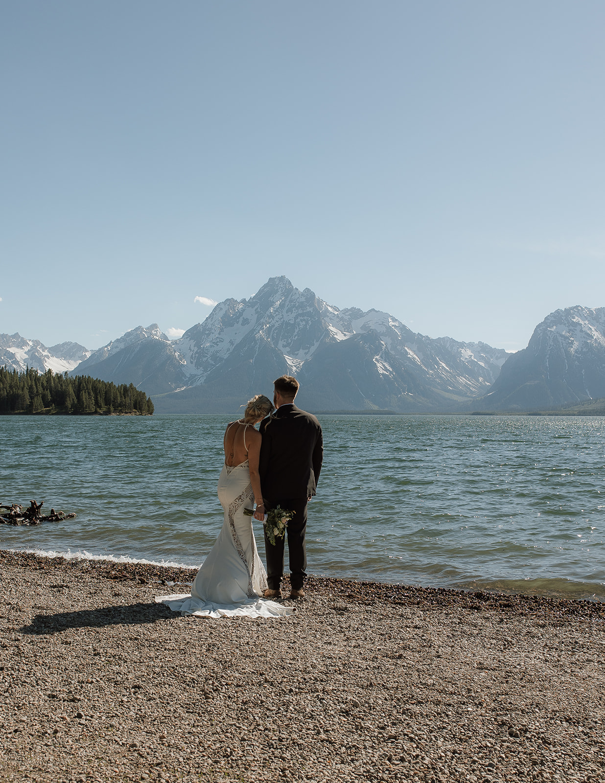 A couple dressed in formal attire stands together on a pebble shore in front of a lake, with snow-capped mountains in the background under a clear blue sky.