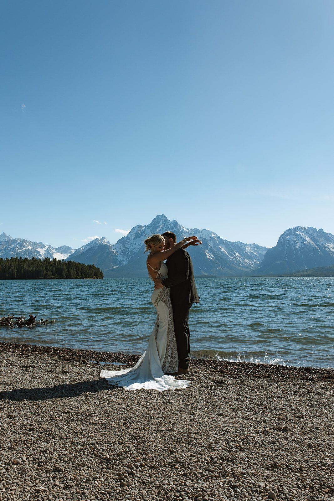 A bride and groom stand by a lake with mountains in the background, embracing each other on a sunny day.