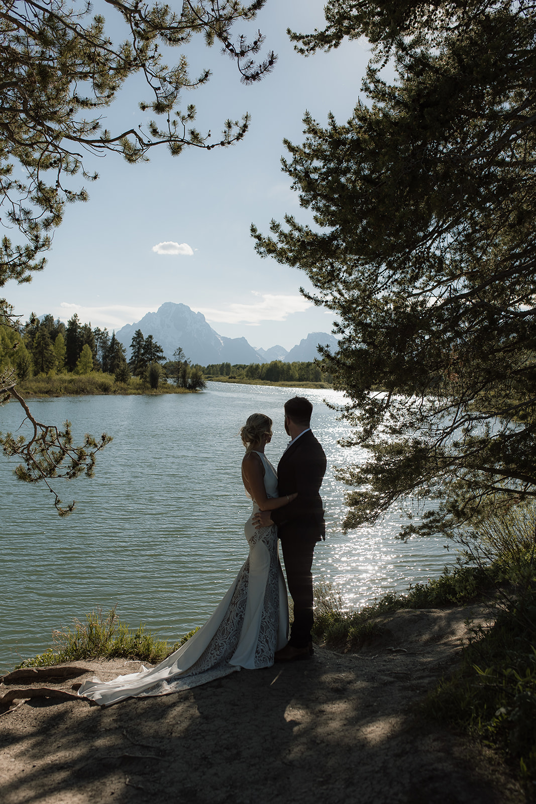 A couple stands by a lake with trees on either side, silhouetted against the water and mountains in the background.