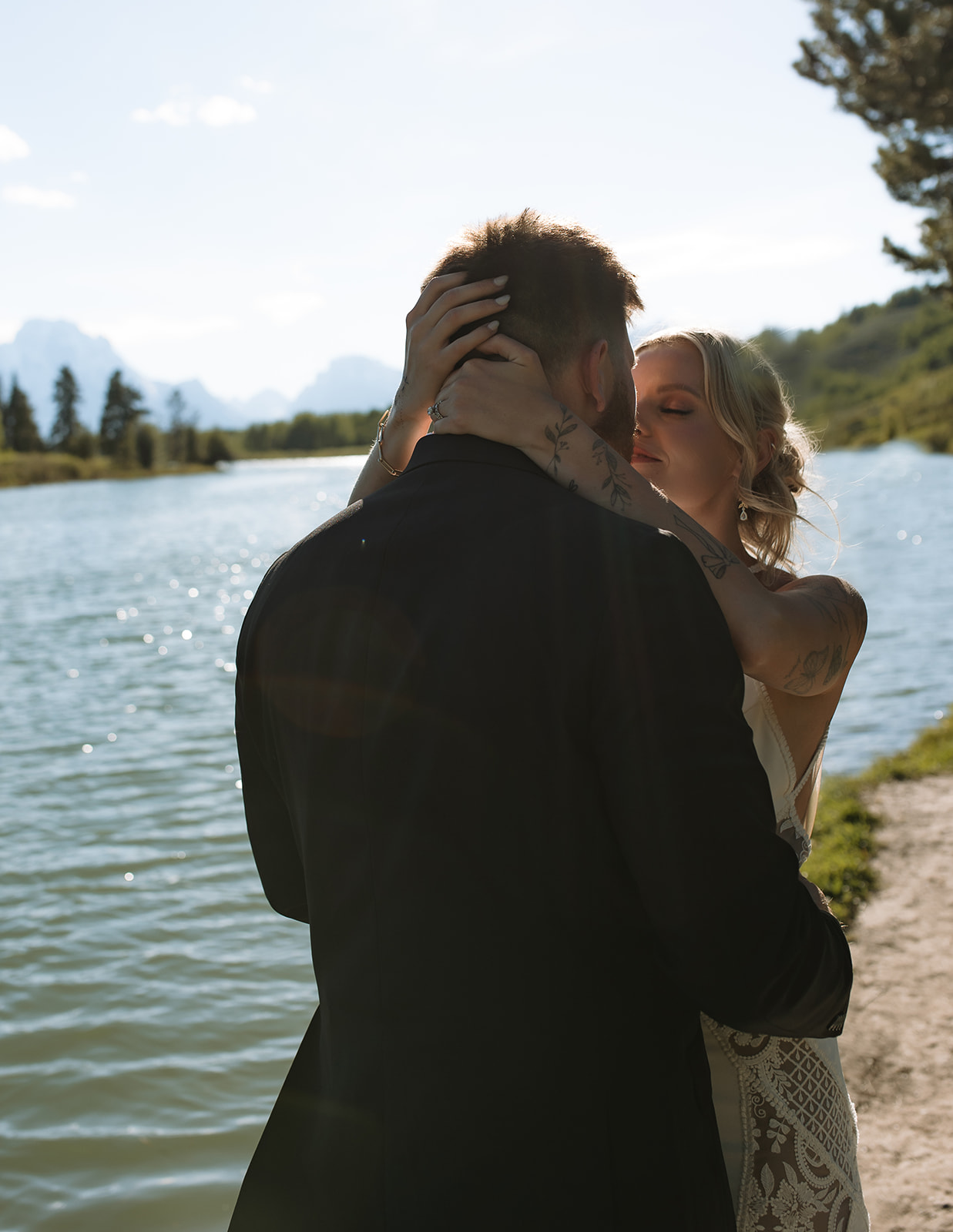 A couple stands by a lake with trees on either side, silhouetted against the water and mountains in the background.