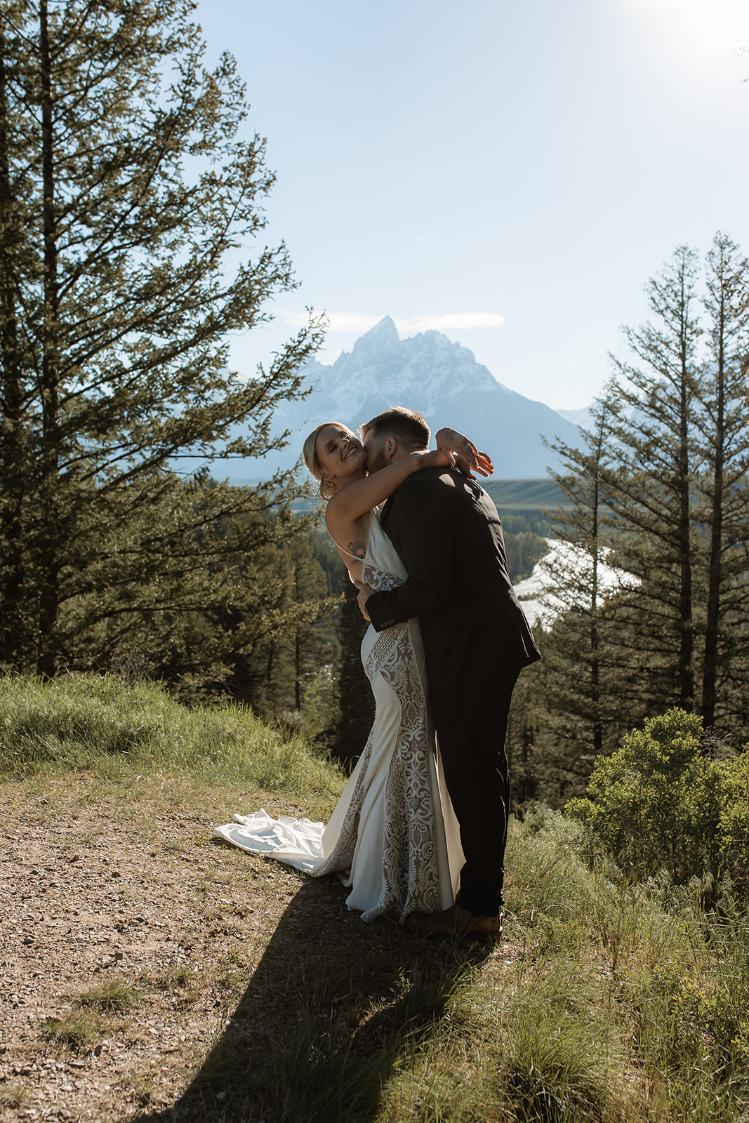 A couple stands by a lake with trees on either side, silhouetted against the water and mountains in the background.