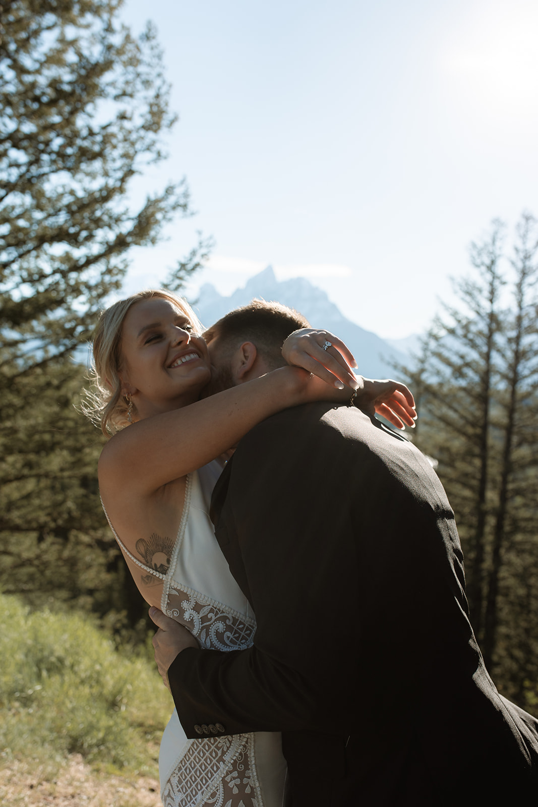 A couple embracing outdoors with a mountain and forest background. The woman is wearing a white dress and the man is in a dark suit.