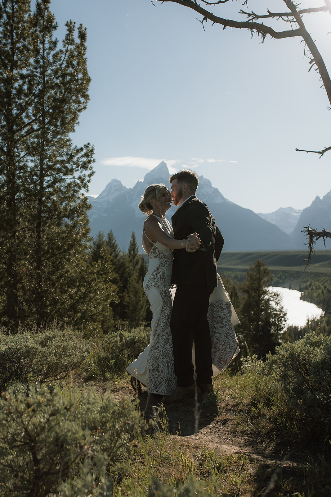 A couple stands by a lake with trees on either side, silhouetted against the water and mountains in the background.