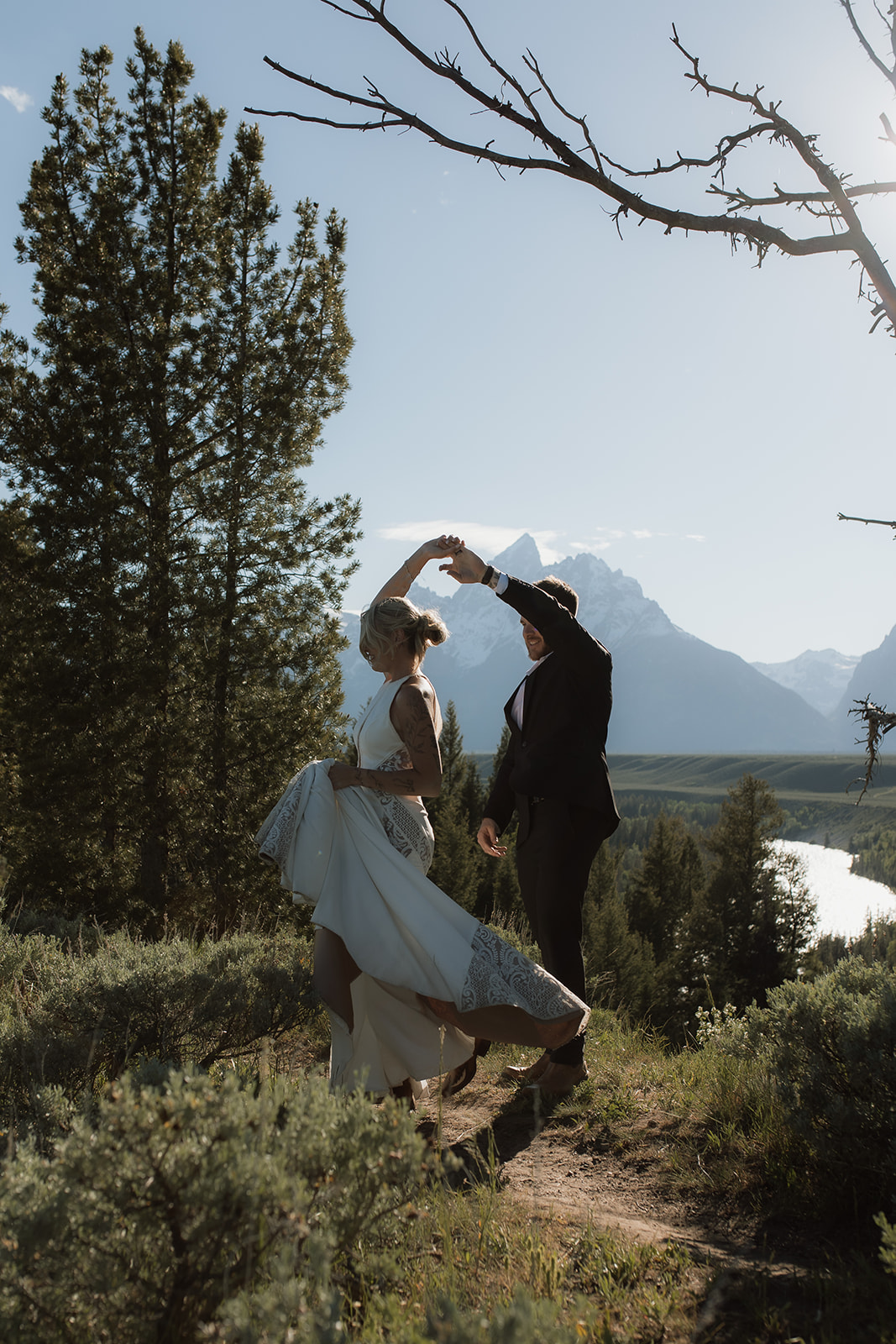 A person in a white dress and another person in a black suit are dancing outdoors on a dirt path, surrounded by trees and mountains in the background for their elopement at jackson hole