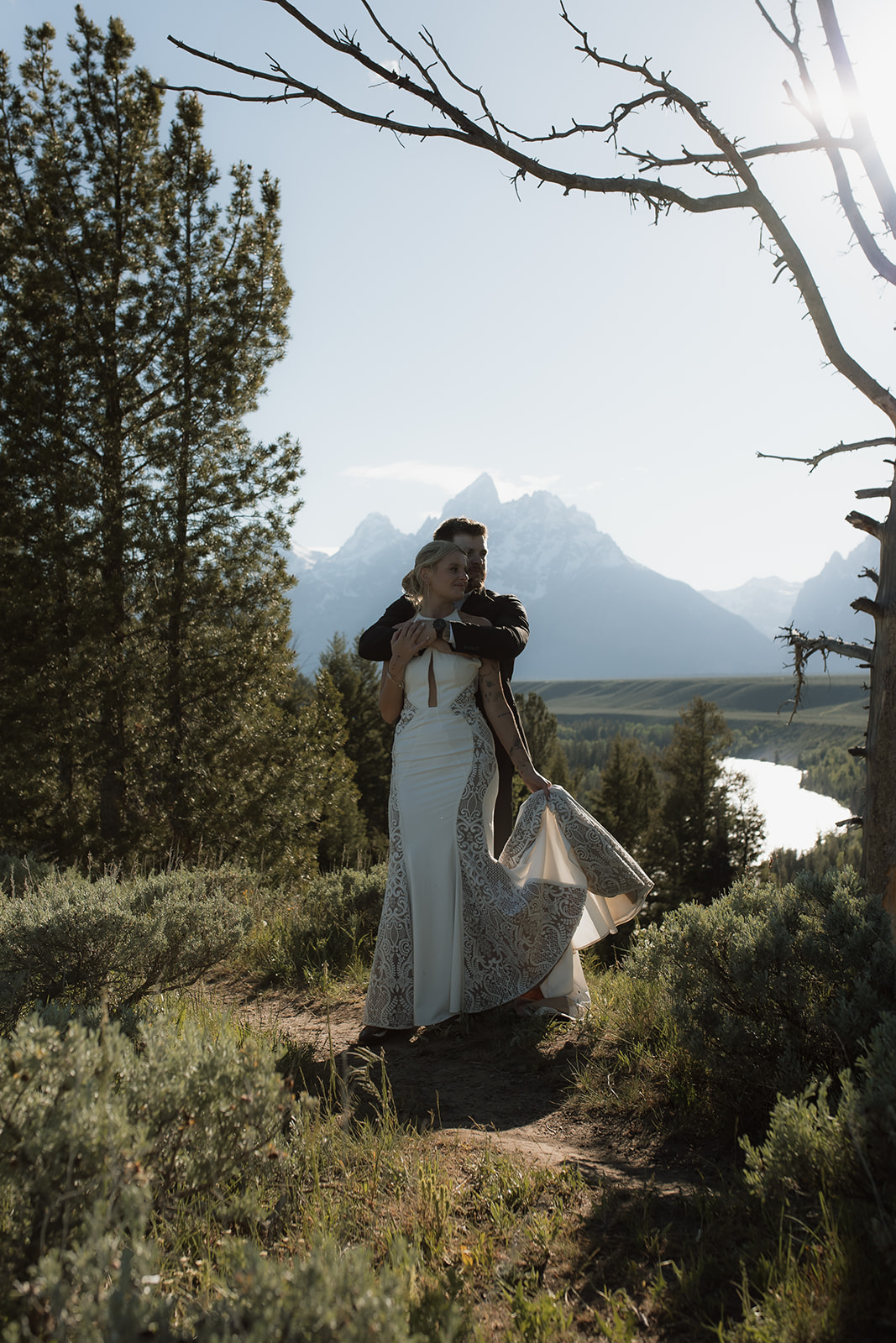 A person in a white dress and another person in a black suit are dancing outdoors on a dirt path, surrounded by trees and mountains in the background for their elopement at jackson hole