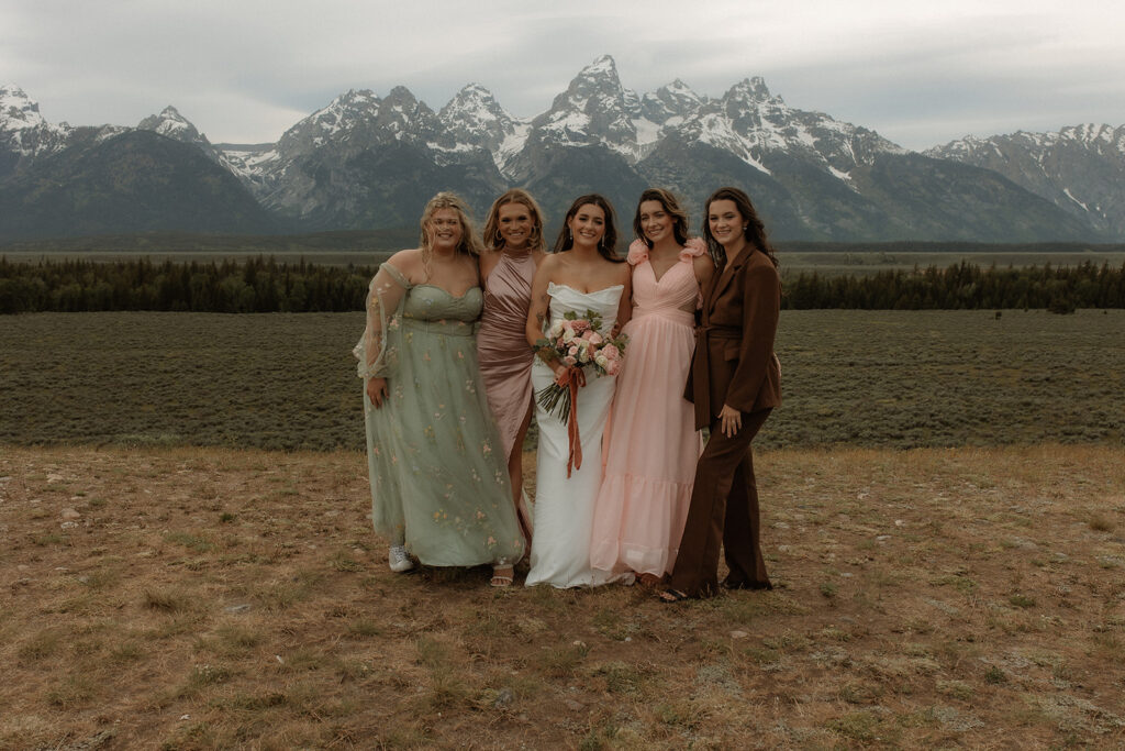 A man, two women, and a younger man stand together outdoors with mountains in the background. The younger woman wears a white dress and holds a bouquet; the older woman is in a pink dress for their elopement at Glacier View turnout