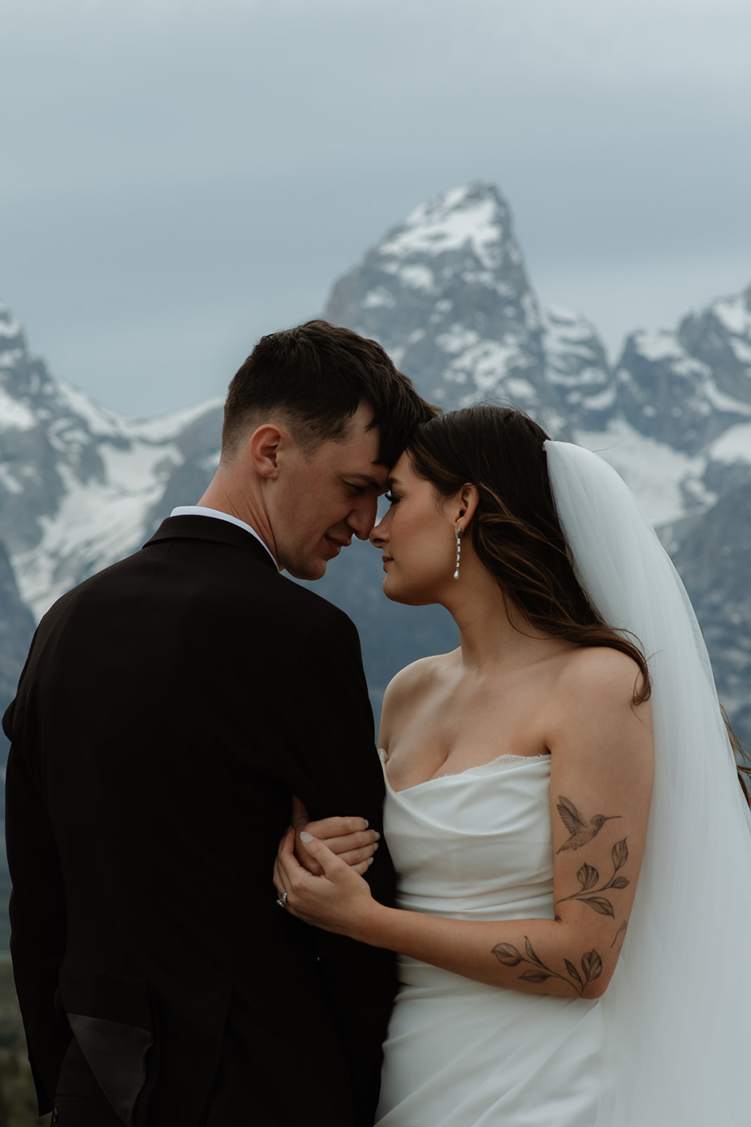 A bride and groom stand close together outdoors, with snowy mountains visible in the background. The bride is wearing a white strapless dress and veil, and has tattoos on her arm.