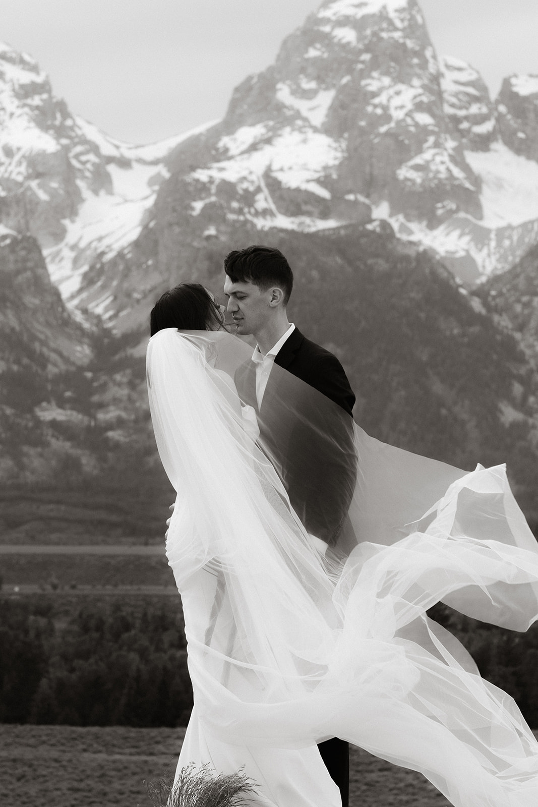 A bride and groom stand close together outdoors, with snowy mountains visible in the background. The bride is wearing a white strapless dress and veil, and has tattoos on her arm.