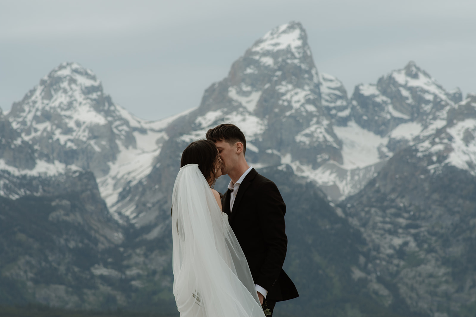 A bride and groom stand close together outdoors, with snowy mountains visible in the background. The bride is wearing a white strapless dress and veil, and has tattoos on her arm.