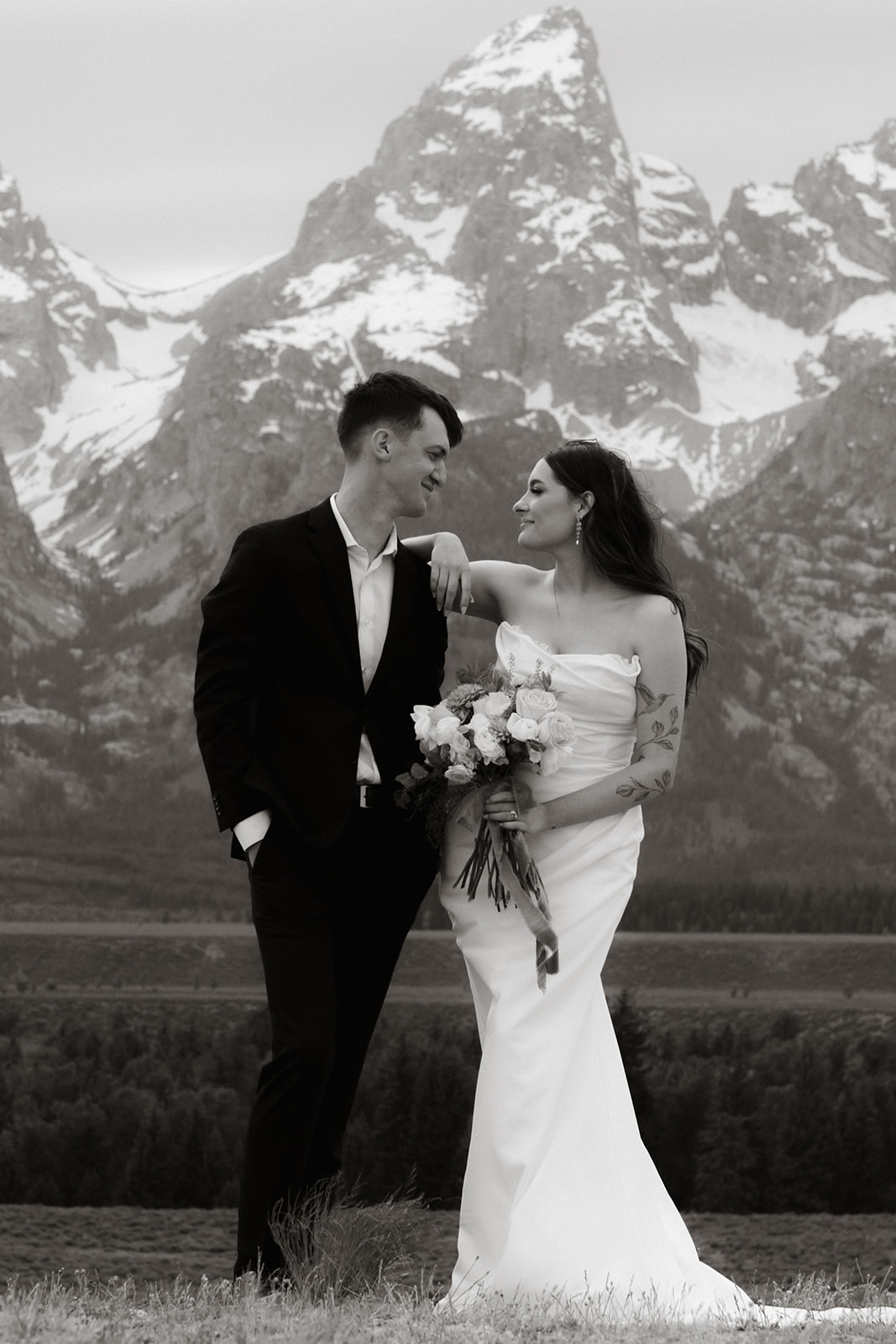 A bride and groom stand close together outdoors, with snowy mountains visible in the background. The bride is wearing a white strapless dress and veil, and has tattoos on her arm.