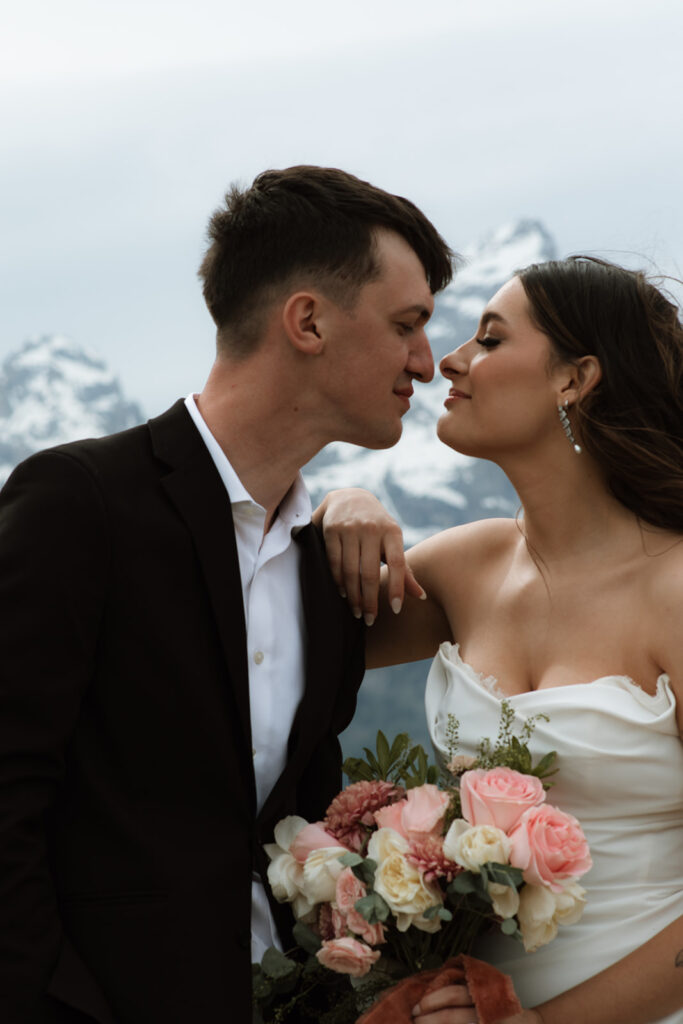 A bride and groom stand close together outdoors, with snowy mountains visible in the background. The bride is wearing a white strapless dress and veil, and has tattoos on her arm.