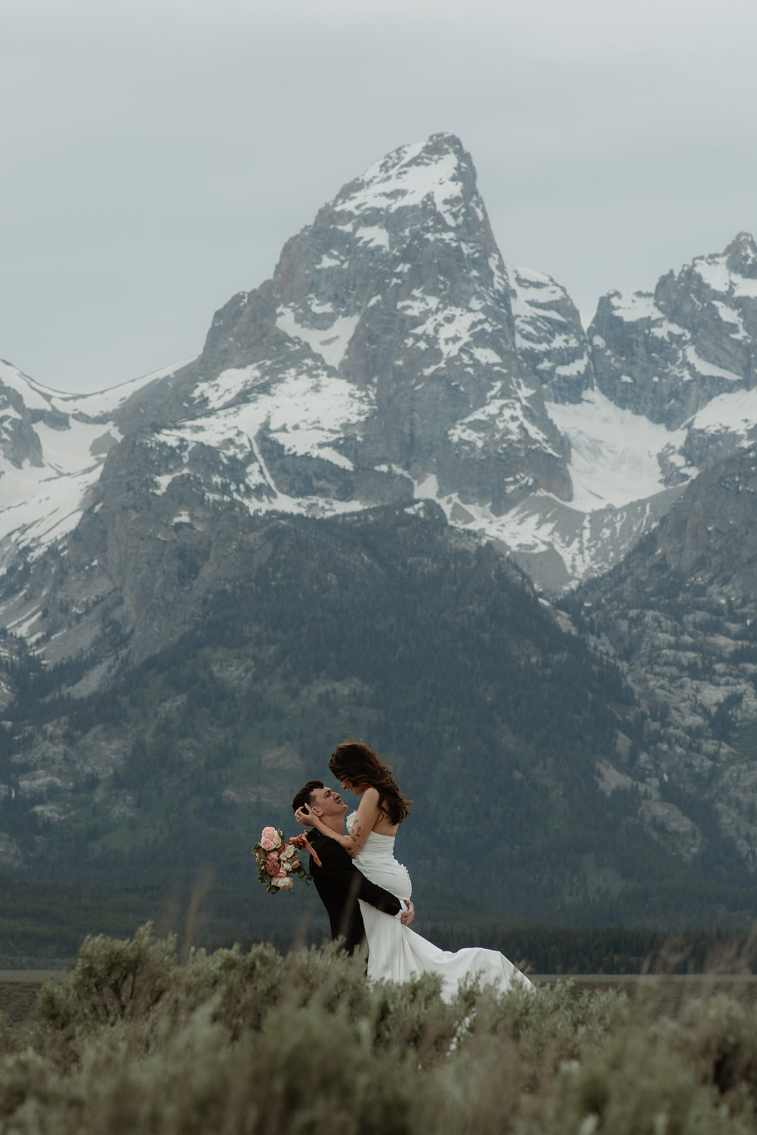 A bride and groom stand close together outdoors, with snowy mountains visible in the background. The bride is wearing a white strapless dress and veil, and has tattoos on her arm.