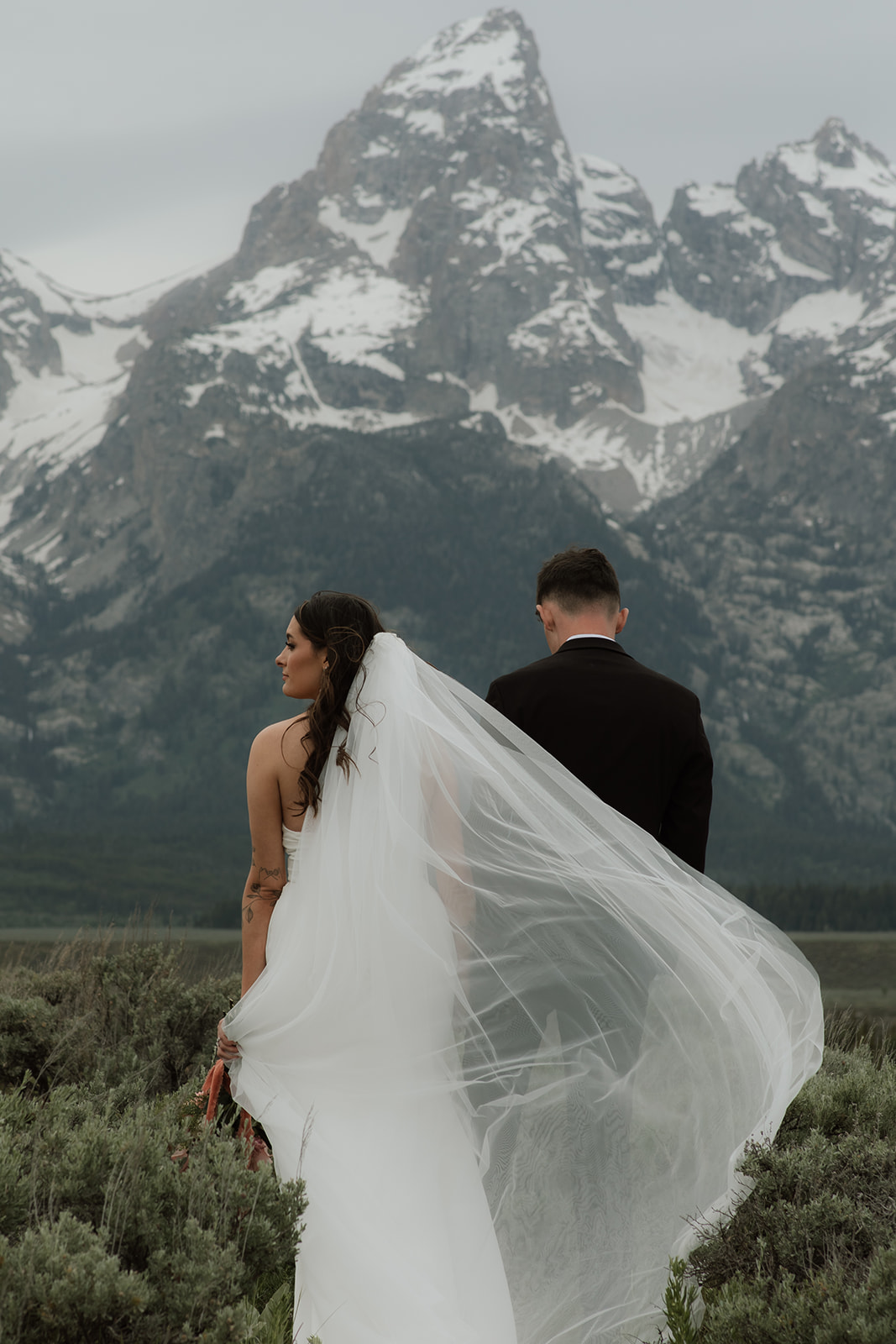 A bride and groom stand close together outdoors, with snowy mountains visible in the background. The bride is wearing a white strapless dress and veil, and has tattoos on her arm.