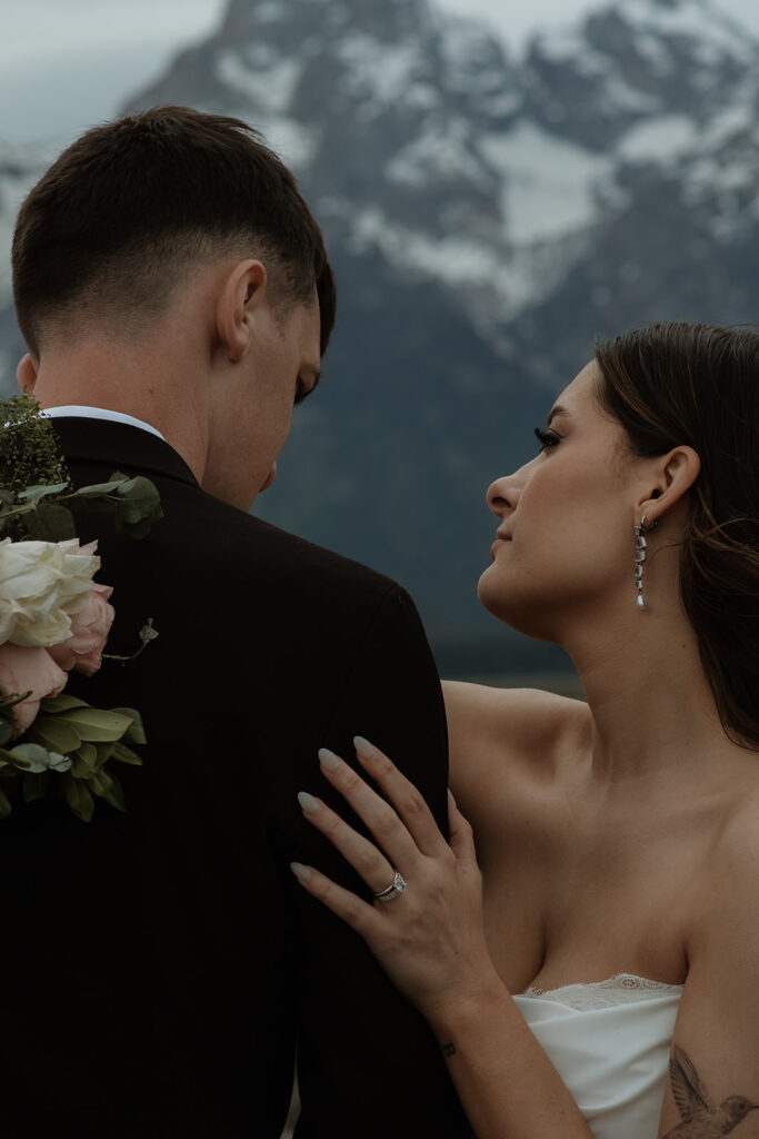 A bride and groom stand close together outdoors, with snowy mountains visible in the background. The bride is wearing a white strapless dress and veil, and has tattoos on her arm.