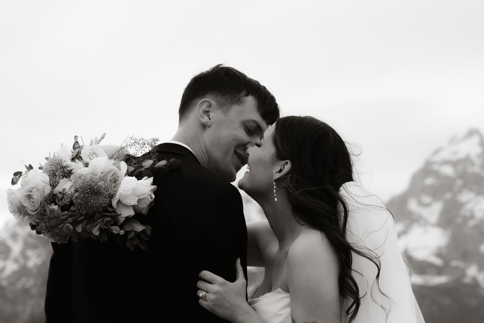 A bride and groom stand close together outdoors, with snowy mountains visible in the background. The bride is wearing a white strapless dress and veil, and has tattoos on her arm.