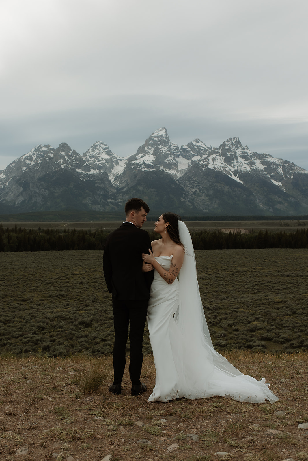 A bride and groom stand close together outdoors, with snowy mountains visible in the background. The bride is wearing a white strapless dress and veil, and has tattoos on her arm.