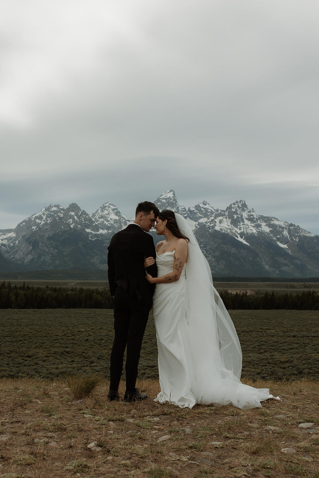 A bride and groom stand close together outdoors, with snowy mountains visible in the background. The bride is wearing a white strapless dress and veil, and has tattoos on her arm.