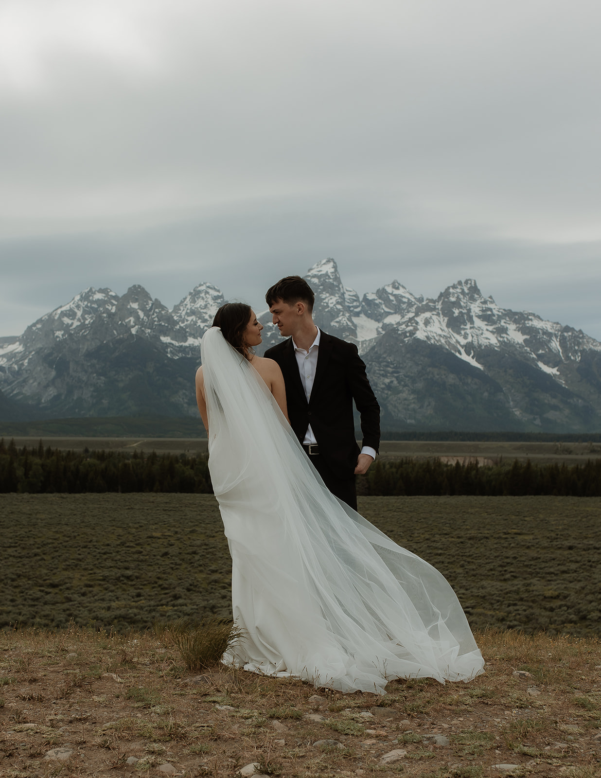 A bride and groom stand close together outdoors, with snowy mountains visible in the background. The bride is wearing a white strapless dress and veil, and has tattoos on her arm.