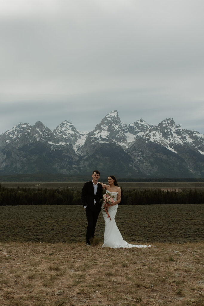 A bride and groom stand close together outdoors, with snowy mountains visible in the background. The bride is wearing a white strapless dress and veil, and has tattoos on her arm.