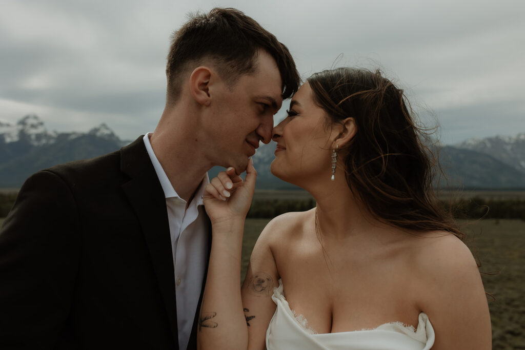 A bride and groom stand close together outdoors, with snowy mountains visible in the background. The bride is wearing a white strapless dress and veil, and has tattoos on her arm.