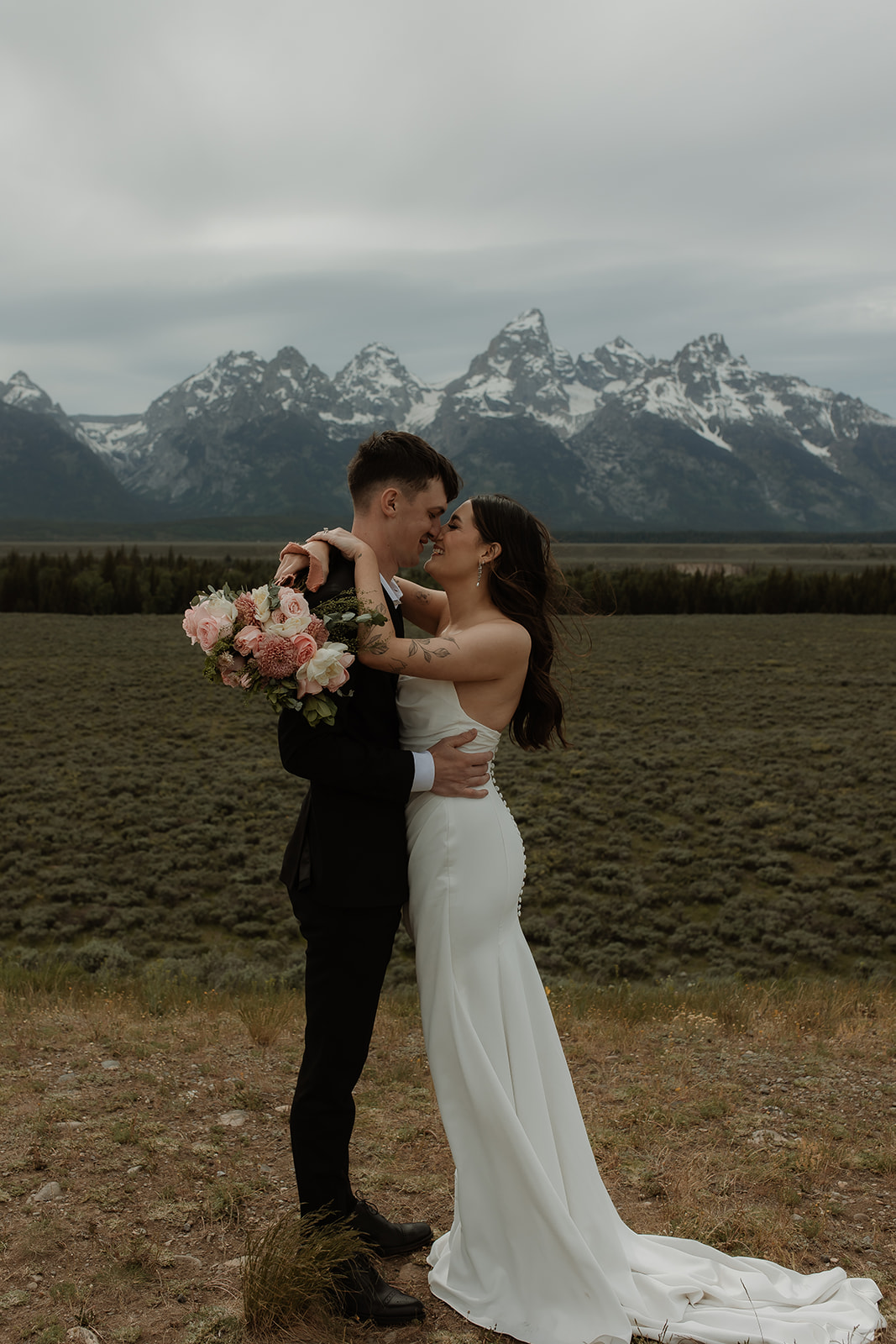 A bride and groom stand close together outdoors, with snowy mountains visible in the background. The bride is wearing a white strapless dress and veil, and has tattoos on her arm.