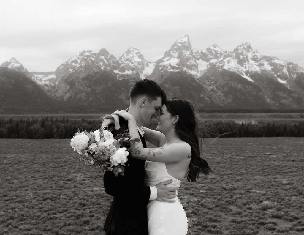 A bride and groom stand close together outdoors, with snowy mountains visible in the background. The bride is wearing a white strapless dress and veil, and has tattoos on her arm.