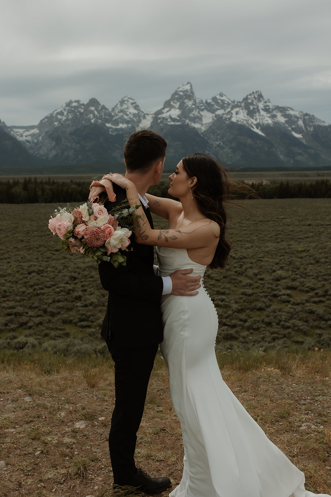 A bride and groom stand close together outdoors, with snowy mountains visible in the background. The bride is wearing a white strapless dress and veil, and has tattoos on her arm.