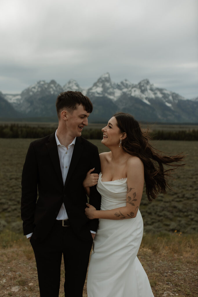 A bride and groom stand close together outdoors, with snowy mountains visible in the background. The bride is wearing a white strapless dress and veil, and has tattoos on her arm.