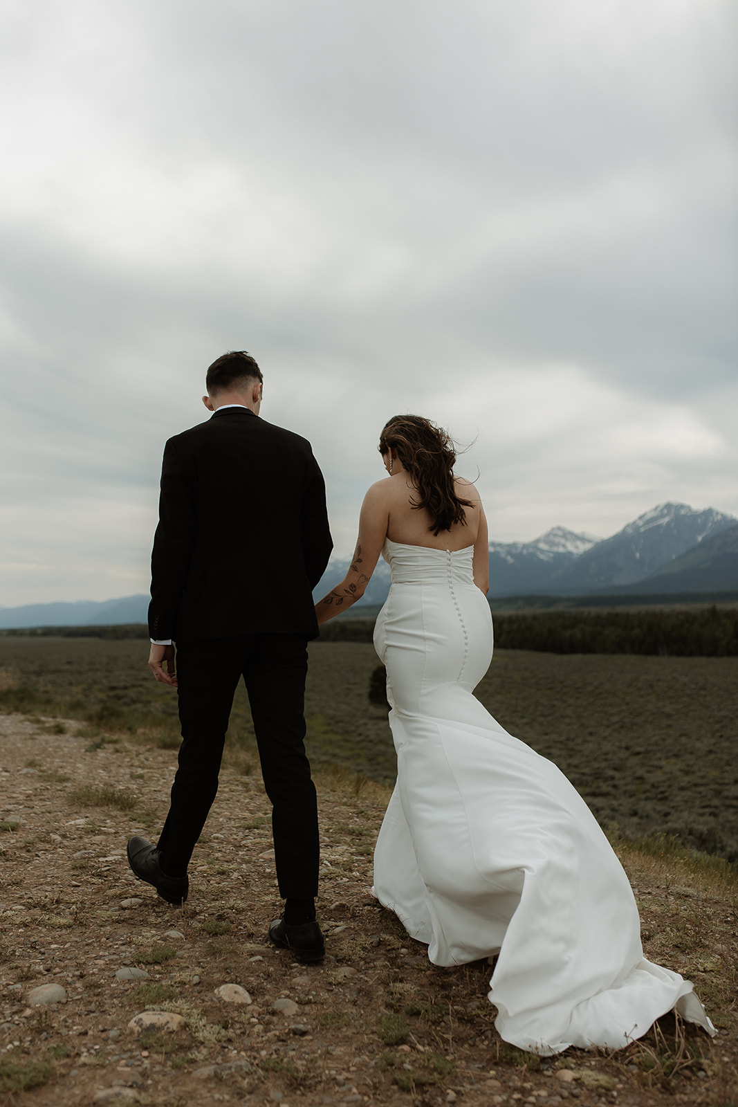 A bride and groom stand close together outdoors, with snowy mountains visible in the background. The bride is wearing a white strapless dress and veil, and has tattoos on her arm.