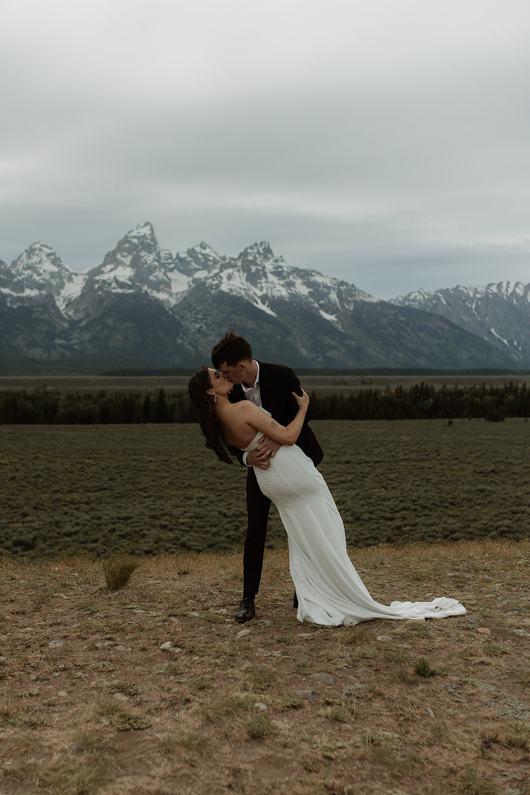 A bride and groom stand close together outdoors, with snowy mountains visible in the background. The bride is wearing a white strapless dress and veil, and has tattoos on her arm.