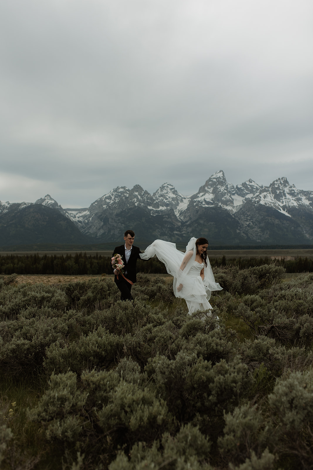 A bride and groom stand close together outdoors, with snowy mountains visible in the background. The bride is wearing a white strapless dress and veil, and has tattoos on her arm.