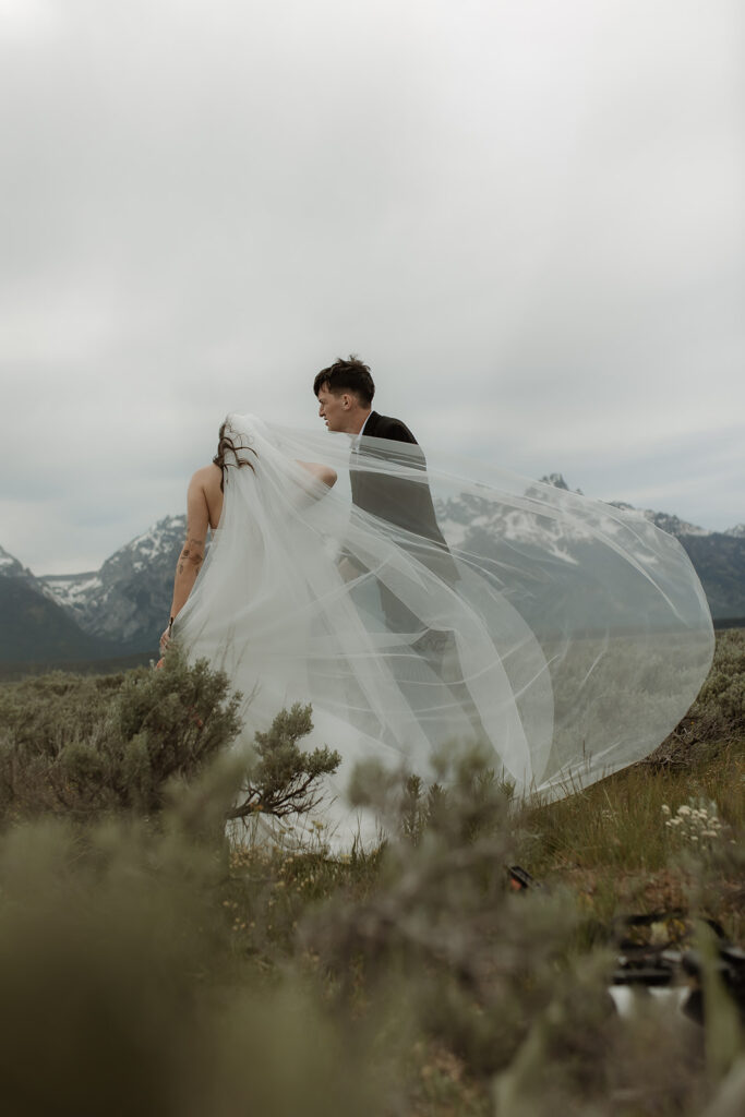 A bride and groom stand close together outdoors, with snowy mountains visible in the background. The bride is wearing a white strapless dress and veil, and has tattoos on her arm.