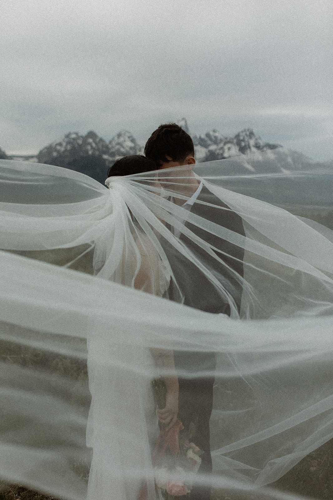 A bride and groom stand close together outdoors, with snowy mountains visible in the background. The bride is wearing a white strapless dress and veil, and has tattoos on her arm.