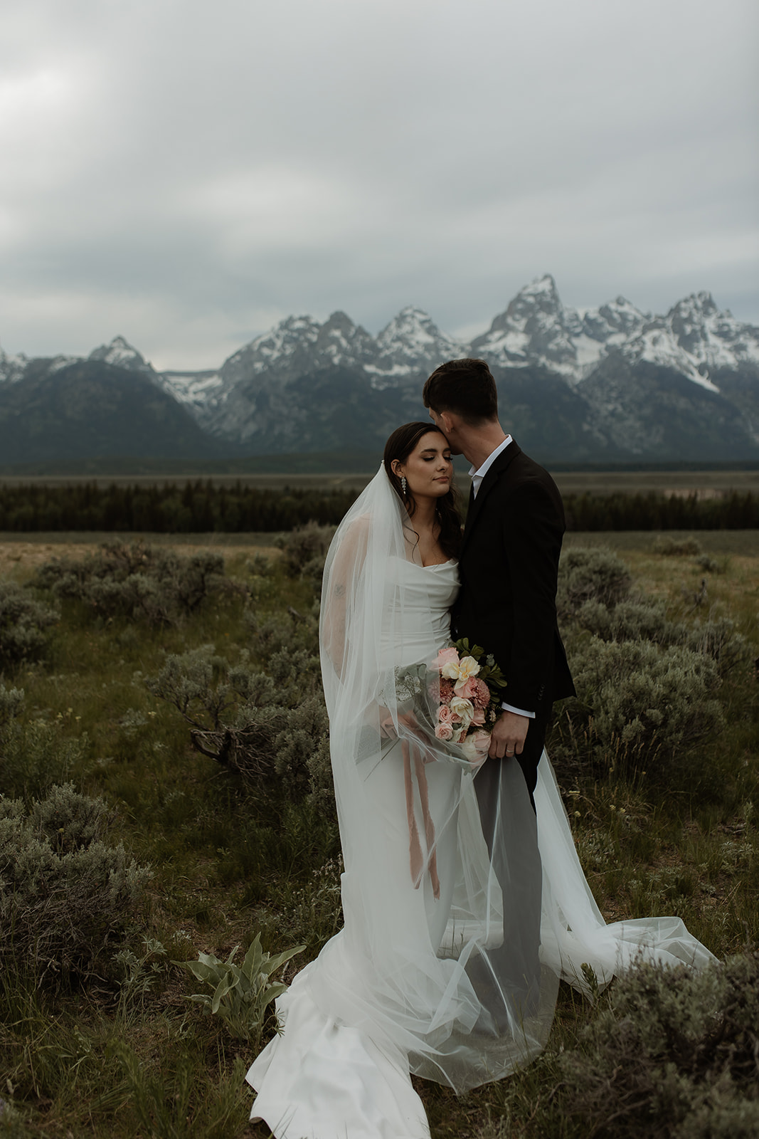 A bride and groom stand close together outdoors, with snowy mountains visible in the background. The bride is wearing a white strapless dress and veil, and has tattoos on her arm.