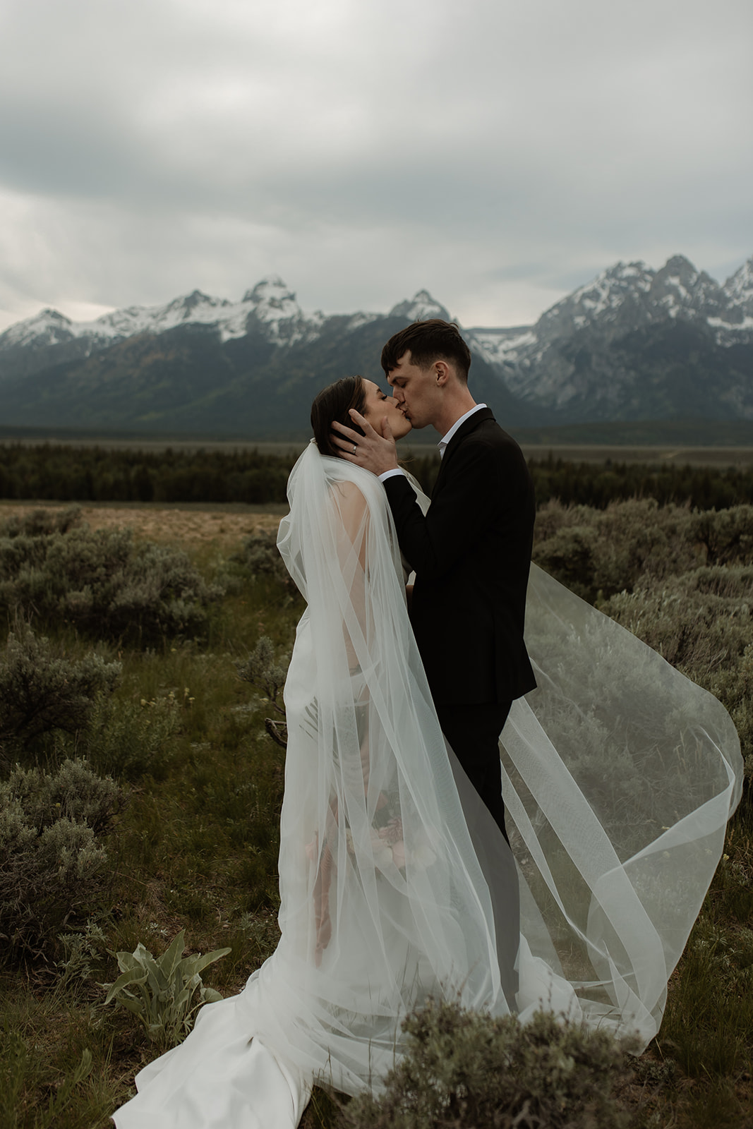 A bride and groom stand close together outdoors, with snowy mountains visible in the background. The bride is wearing a white strapless dress and veil, and has tattoos on her arm.