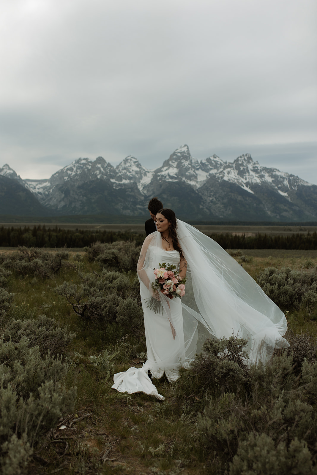 A bride and groom stand close together outdoors, with snowy mountains visible in the background. The bride is wearing a white strapless dress and veil, and has tattoos on her arm.