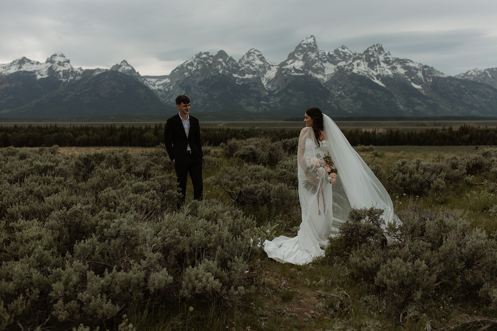 A bride and groom stand close together outdoors, with snowy mountains visible in the background. The bride is wearing a white strapless dress and veil, and has tattoos on her arm.