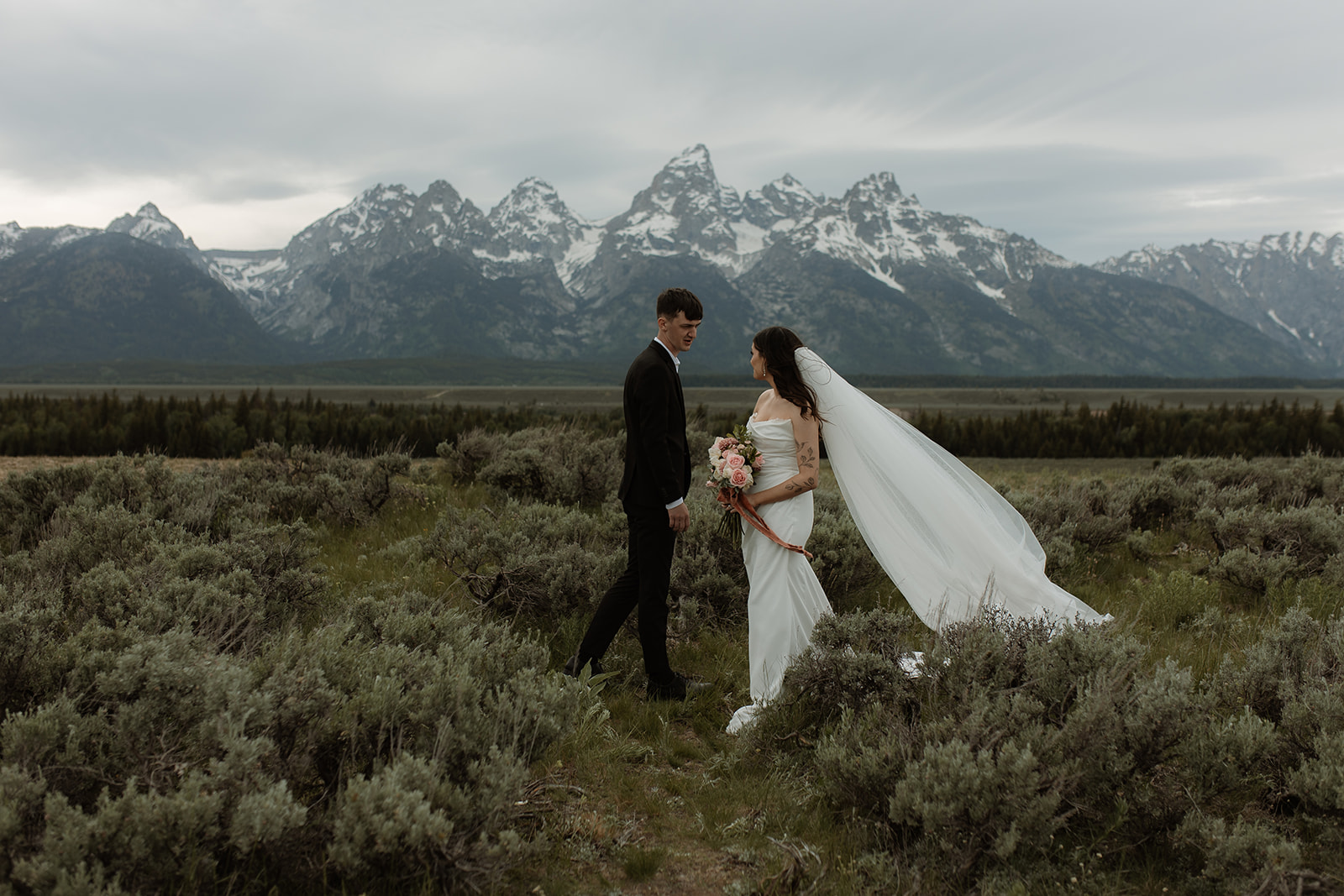 A bride and groom stand together in front of a mountainous landscape. The bride holds a bouquet of flowers and wears a white wedding dress with a veil, while the groom wears a dark suit for their elopement at Glacier view turnout