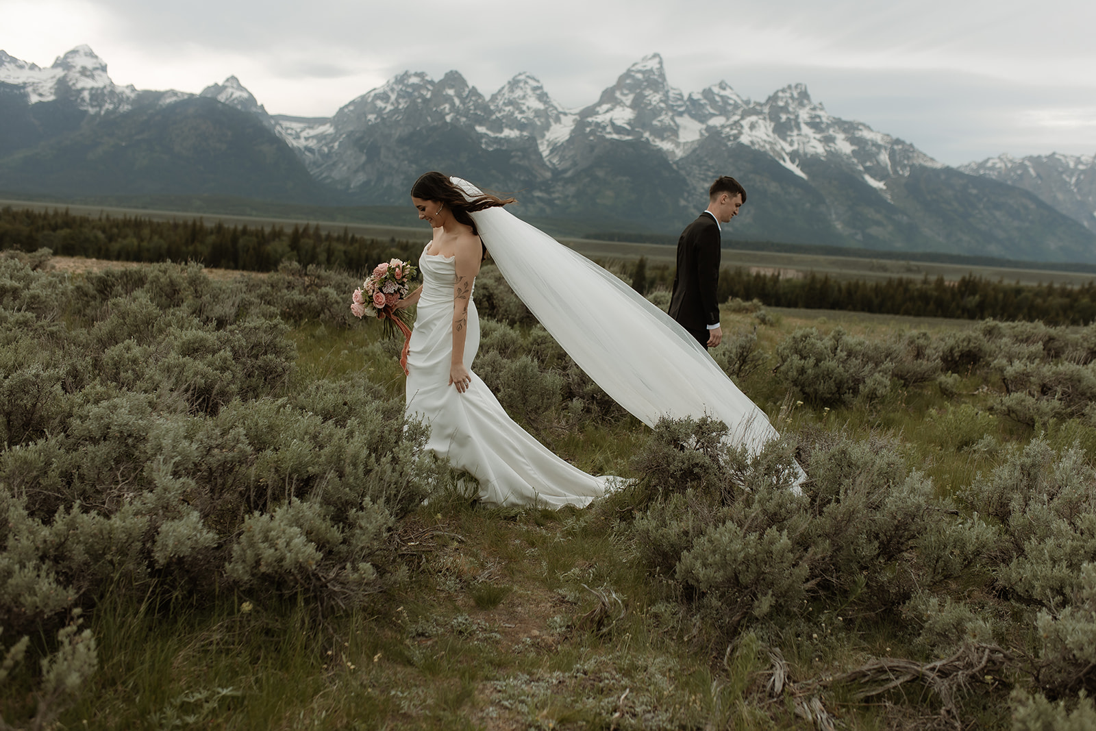 A bride and groom stand together in front of a mountainous landscape. The bride holds a bouquet of flowers and wears a white wedding dress with a veil, while the groom wears a dark suit for their elopement at Glacier view turnout