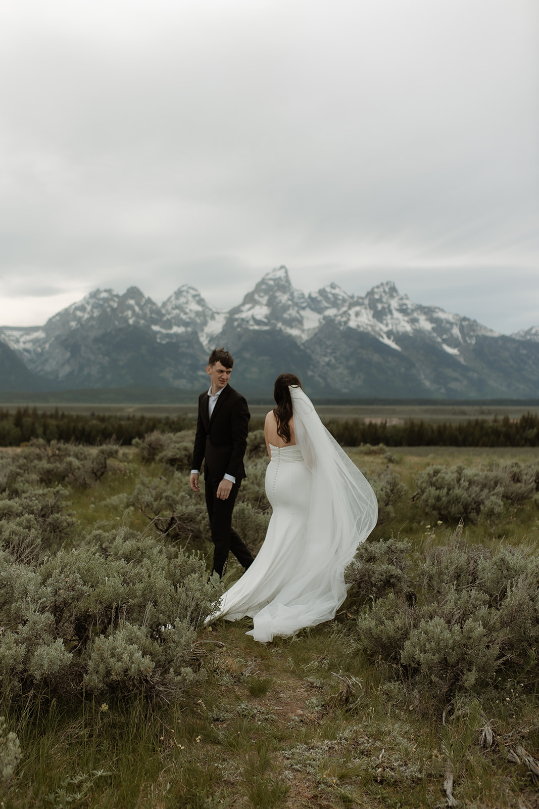 A bride and groom stand together in front of a mountainous landscape. The bride holds a bouquet of flowers and wears a white wedding dress with a veil, while the groom wears a dark suit for their elopement at Glacier view turnout