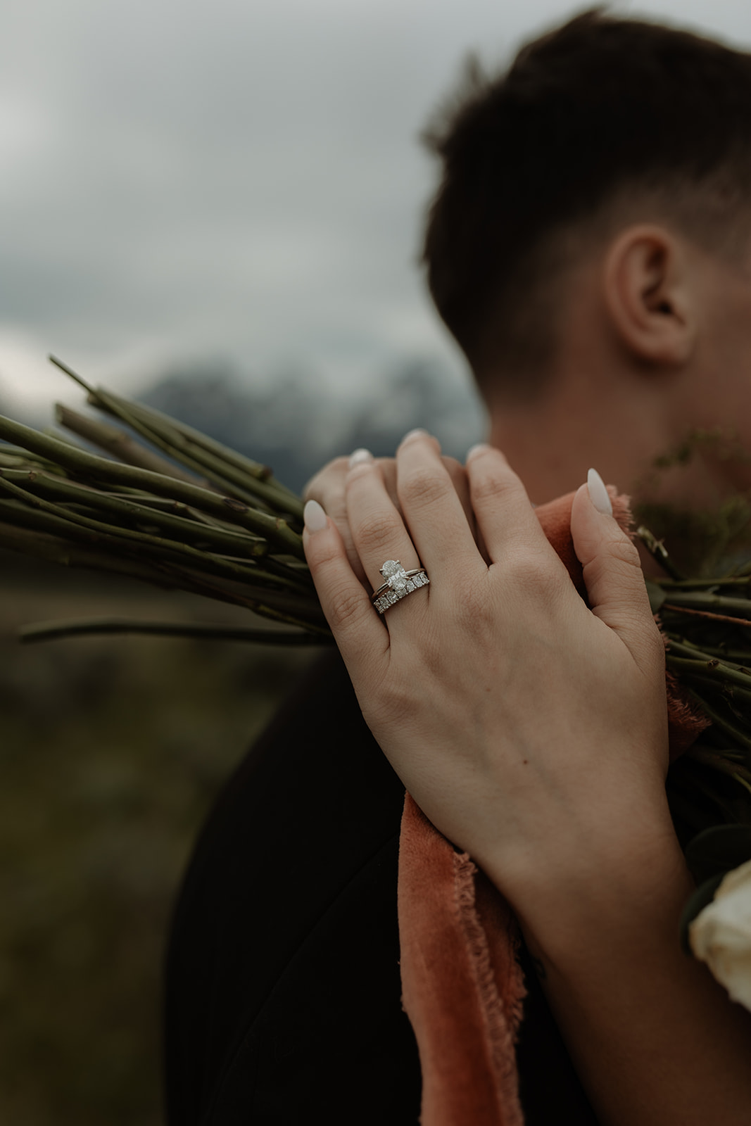 A bride and groom stand together in front of a mountainous landscape. The bride holds a bouquet of flowers and wears a white wedding dress with a veil, while the groom wears a dark suit for their elopement at Glacier view turnout  