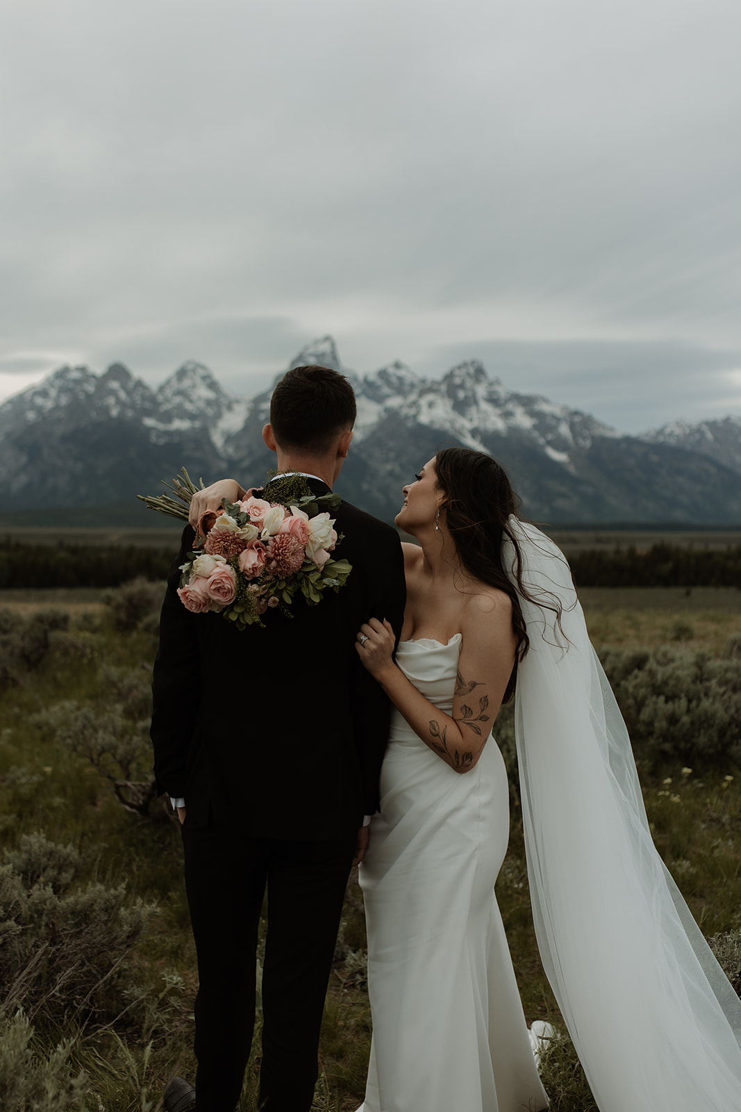 A bride and groom stand together in front of a mountainous landscape. The bride holds a bouquet of flowers and wears a white wedding dress with a veil, while the groom wears a dark suit for their elopement at Glacier view turnout  