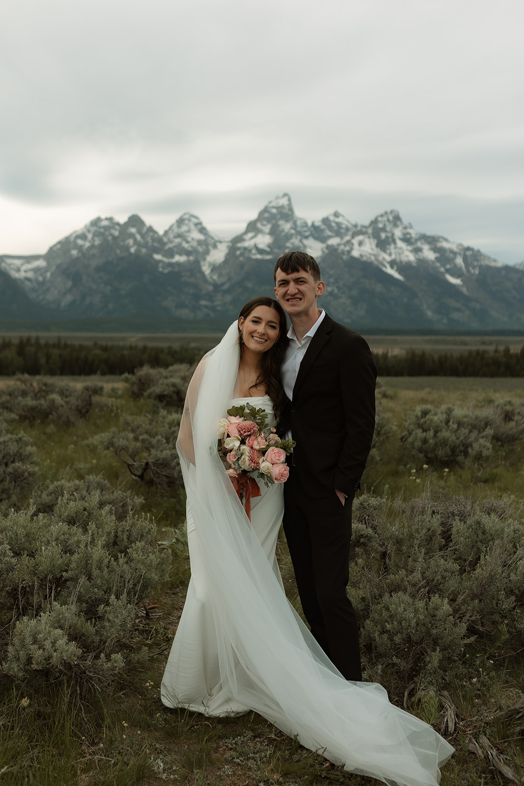 A bride and groom stand together in front of a mountainous landscape. The bride holds a bouquet of flowers and wears a white wedding dress with a veil, while the groom wears a dark suit for their elopement at Glacier view turnout