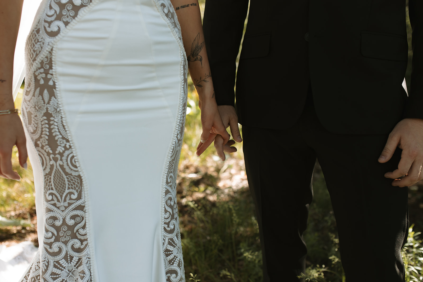 A couple holds hands outdoors. The woman wears a white lace-trimmed dress, and the man is dressed in a black suit.