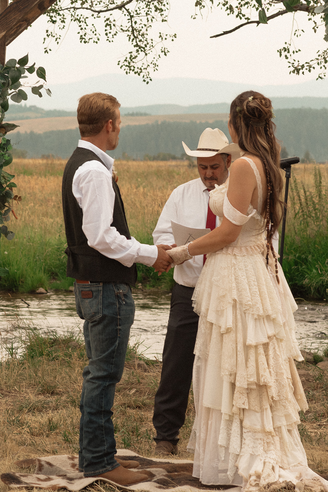 A couple stands under a wooden arch adorned with greenery and flowers, holding hands, while an officiant in a white hat and shirt reads from a book during an outdoor wedding ceremony.