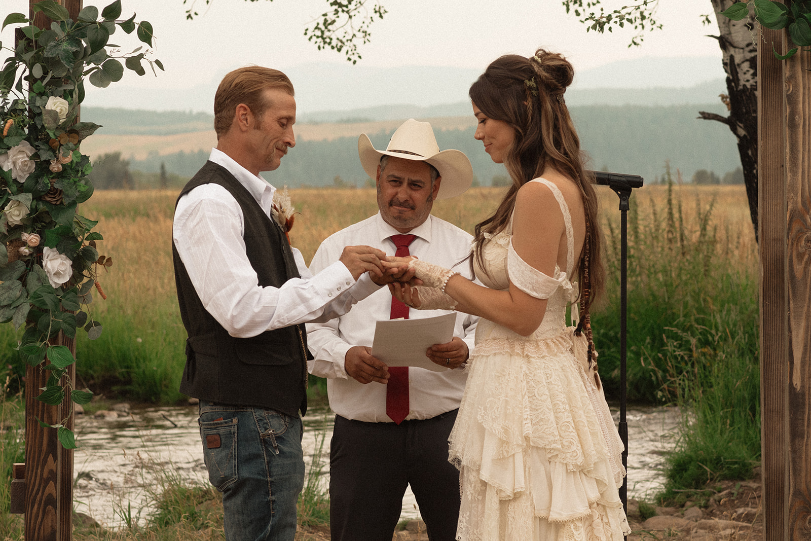 A couple stands under a wooden arch adorned with greenery and flowers, holding hands, while an officiant in a white hat and shirt reads from a book during an outdoor wedding ceremony.